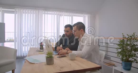 两位成年白种人商人使用笔记本电脑并在办公室室内进行讨论的肖像视频的预览图