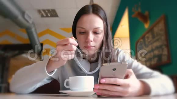 女人正在用勺子喝热巧克力看着坐在咖啡馆里的手机屏幕视频的预览图