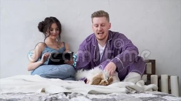 可爱的年轻夫妇相爱坐在床上拿着平板电脑抚摸着一只狗视频的预览图