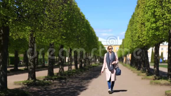一个穿着粉红色外套带着运动鞋和太阳镜的女孩走在公园的小巷里手里拿着一部手机视频的预览图