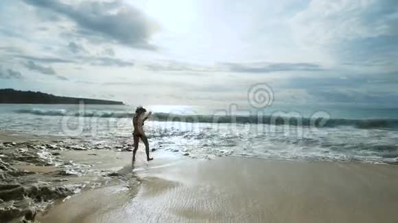 穿着黑色比基尼的快乐可爱女孩在巴厘岛海滨的海滩上跳跃和奔跑年轻女性旅行者视频的预览图
