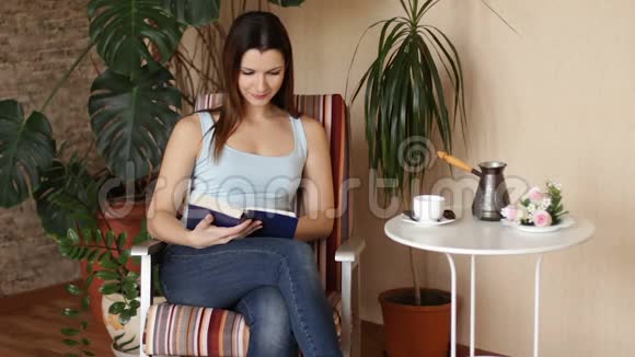 年轻迷人的女人在客厅里坐在舒适的椅子上读有趣的书视频的预览图