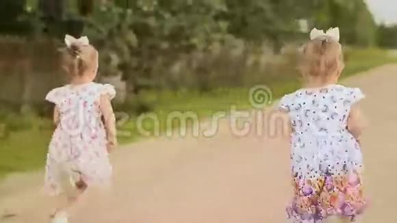 两个小孪生姐妹沿着森林的道路奔跑夏日阳光明媚姑娘们穿着浅色的连衣裙视频的预览图