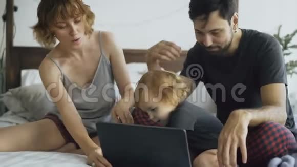 一个年轻的家庭有一个可爱的小女孩和女儿一起玩而她坐在家里的床上打手提电脑视频的预览图