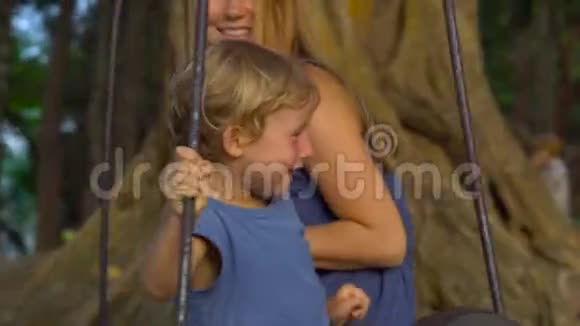 母亲和儿子在热带公园的一棵大热带树上悬挂着一个异国情调的老秋千旅行与儿童概念视频的预览图