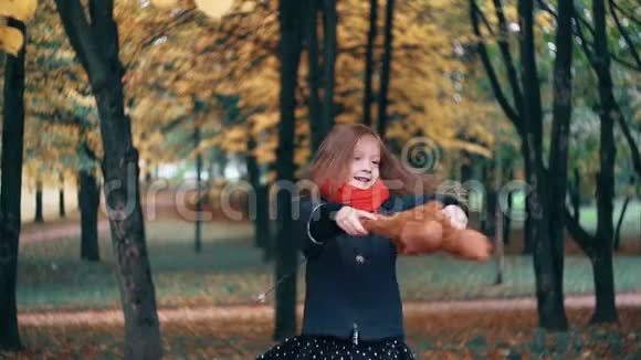 有趣的欢快的可爱的小女孩拿着她的玩具旋转泰迪熊黄色的叶子从树上慢慢地落在它们身上视频的预览图