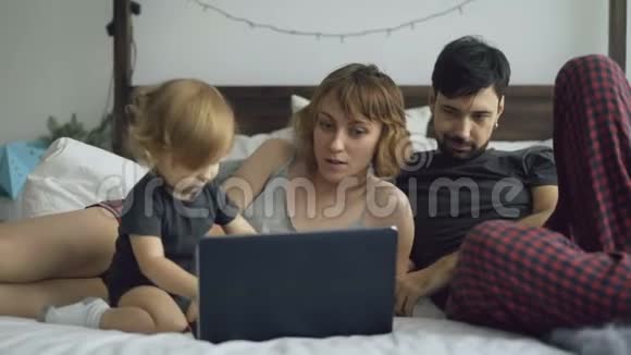 一个年轻的家庭有一个可爱的小女孩和女儿一起玩而她坐在家里的床上打手提电脑视频的预览图
