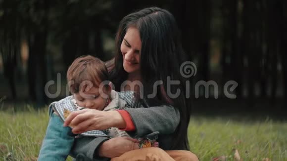 年轻漂亮的妈妈和她可爱的小儿子玩耍小儿子正坐在秋天公园的膝上视频的预览图