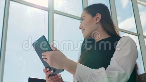 年轻的女乘客在机场一边用平板电脑一边等着她的航班微笑着阳光明媚的一天视频的预览图