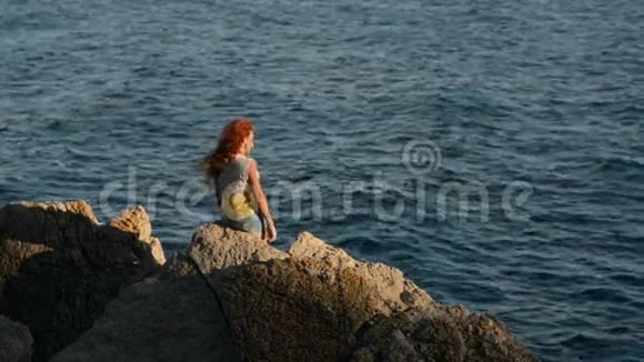一位头发飞扬的年轻女子坐在岩石海岸上视频的预览图