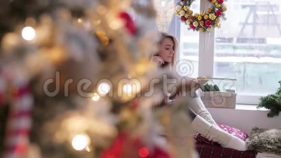 年轻的金发女人穿着紧身裤坐在窗户上读一本书圣诞节内部视频的预览图