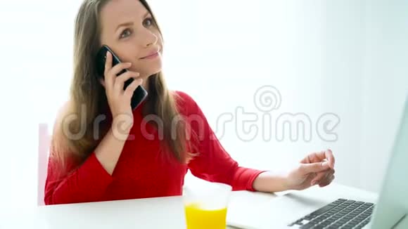 微笑的女人坐在白色的家庭办公室里用智能手机说话视频的预览图