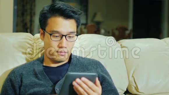 一个带眼镜的年轻正宗亚洲男人他坐在家里的沙发上看电子书视频的预览图
