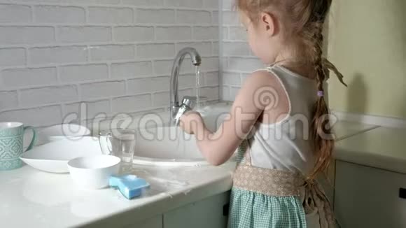 穿着围裙的漂亮小女孩站在明亮厨房的椅子上用水冲洗盘子帮助父母视频的预览图