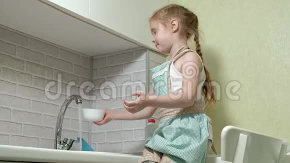 穿着围裙的漂亮小女孩站在明亮厨房的椅子上用水冲洗盘子帮助父母视频的预览图