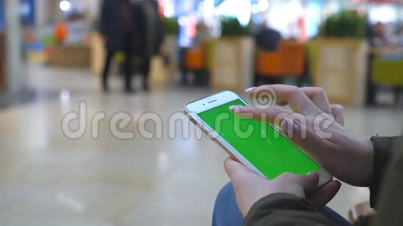 女性手使用现代智能手机与绿色屏幕年轻女孩坐在商场的长椅上轻敲视频的预览图