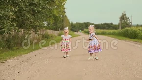 两个穿着白色连衣裙的小双胞胎姐妹在沿着田野的路上一起跑遥远的森林视频的预览图