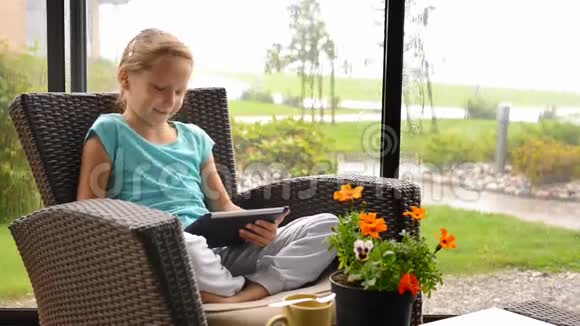 坐在阳台扶手椅上拿着数码平板电脑的小女孩视频的预览图