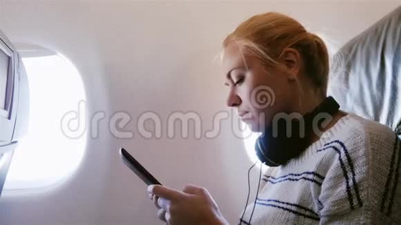 年轻漂亮的女人正在飞机上飞行它使用智能手机听音乐视频的预览图