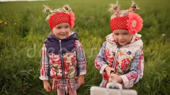 两个可爱的小双胞胎姐妹在绿色的草地上拍照用手机和棍子自拍相互交流视频的预览图