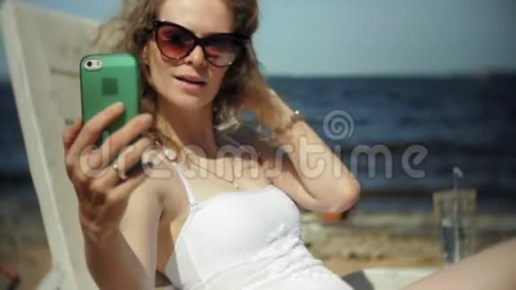 一个穿着白色比基尼的年轻女孩躺在海边沙滩上的休息室里晒太阳并使用智能手机视频的预览图