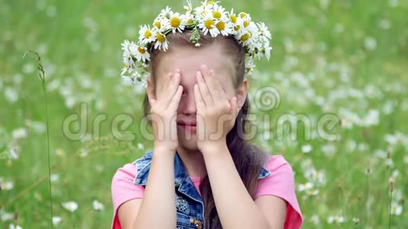 在一片洋甘菊的草坪上一个戴着雏菊花圈的甜美女孩微笑着双手贴在脸颊上视频的预览图