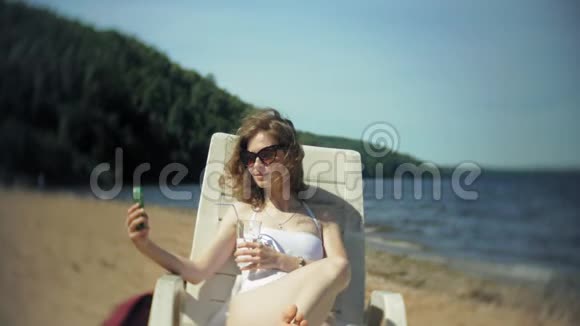 一个穿着白色比基尼的年轻女孩躺在海边沙滩上的休息室里晒太阳并使用智能手机视频的预览图