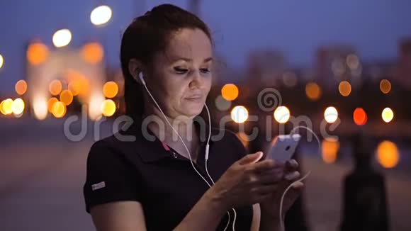 晚上一个女人站在河堤上用手指滚动她智能手机的屏幕视频的预览图