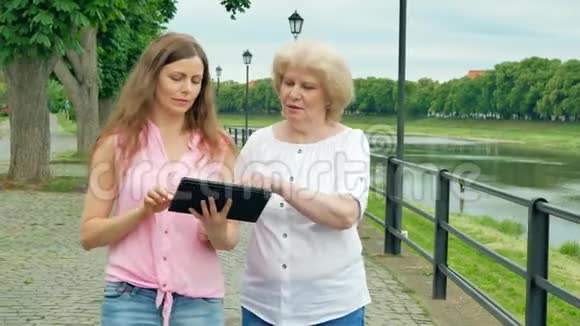 幸福的老年妇女和年轻妇女沿着海滨和平板电脑散步河流的背景视频的预览图