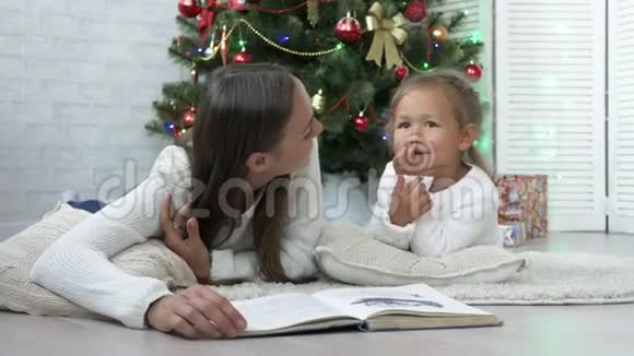 母亲带着女儿躺在地板上在圣诞树附近看书视频的预览图