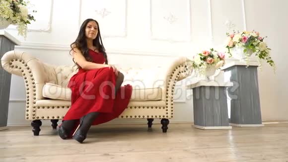 年轻漂亮的黑发女孩穿着红裙子坐在美容院的沙发上视频的预览图