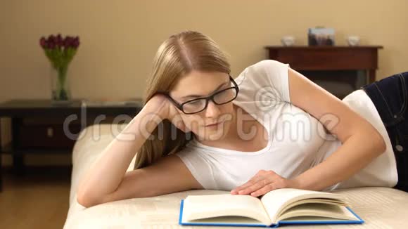 美丽的年轻女子穿着白色的t恤和眼镜轻松地躺在沙发上享受着看书的乐趣视频的预览图