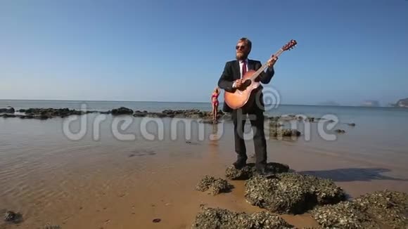 吉他手在沙滩上为金发女孩在岩石上熟练地演奏视频的预览图