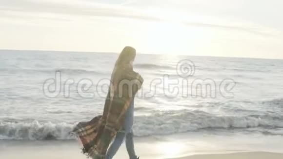 年轻女子走在海边在寒冷的风天穿着格子花衣的女性独自在海滩上度过视频的预览图