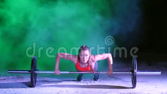 运动女孩正在用杠铃做俯卧撑晚上在轻烟雾中在五彩探照灯下在视频的预览图