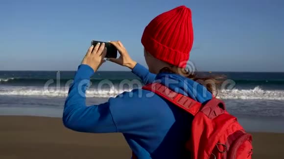 活跃的徒步旅行者女人在海滩上散步在她的智能手机上拍照戴着背包的白人年轻女子视频的预览图