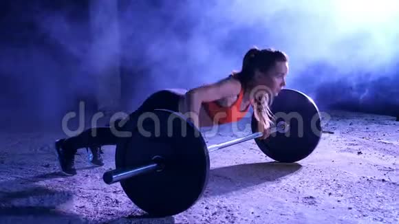 运动女孩正在用杠铃做俯卧撑晚上在轻烟雾中在五彩探照灯下在视频的预览图