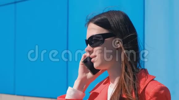 戴墨镜的年轻黑发女人的肖像在蓝色建筑旁边的电话里说话视频的预览图
