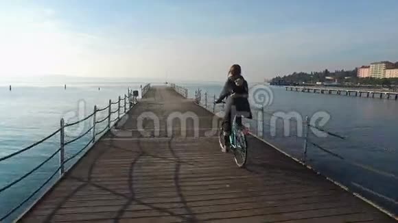 4K在阳光明媚的日子里快乐的女孩骑着自行车在码头上的蓝海上空休假时间视频的预览图