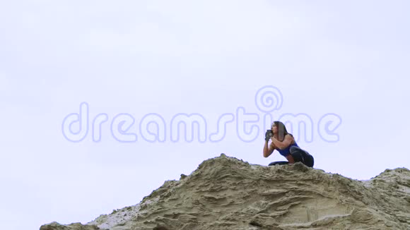 年轻的运动的女人穿着蓝色上衣和条纹腿裤做运动蹲在采石场的沙质顶部健身视频的预览图
