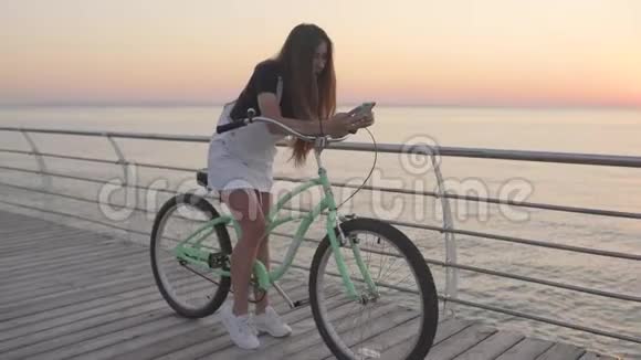年轻漂亮的女人在日出或日落时使用智能手机在海边骑老式自行车视频的预览图