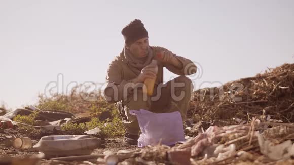 一个肮脏的无家可归的饥饿男人在垃圾堆里喝着包裹里丢失的果汁步行去寻找视频的预览图