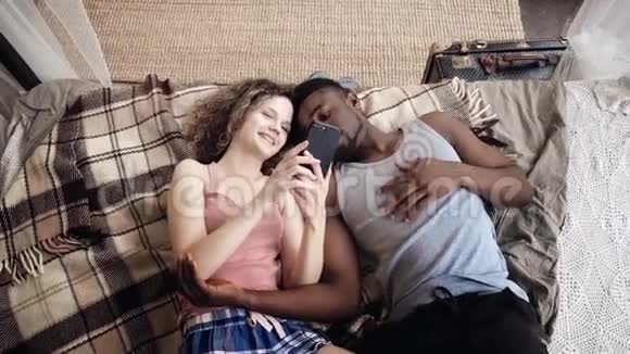 多种族夫妇躺在床上男人和女人一起度过快乐和粉丝的时光使用智能手机上网视频的预览图