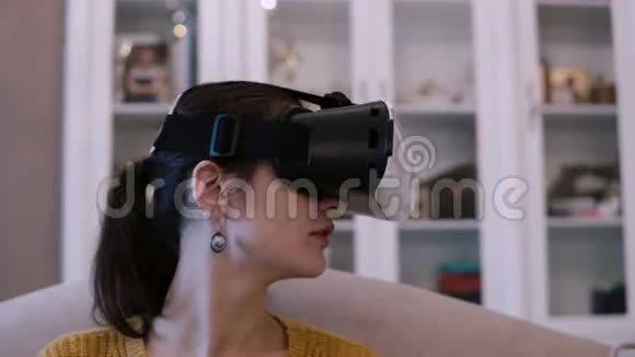 近景的年轻女子在虚拟现实眼镜坐在沙发上的家女性在VR耳机里环顾四周视频的预览图