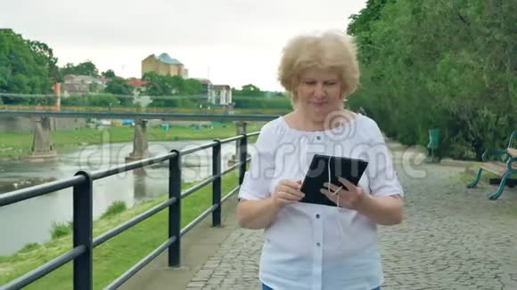 老年妇女沿着海滨散步使用带有耳机的平板电脑河流的背景视频的预览图