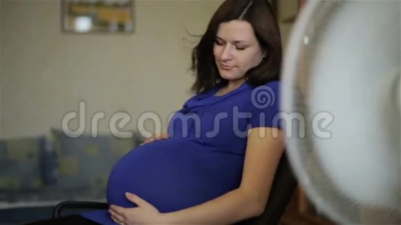 一位穿着蓝色连衣裙的孕妇正坐在风扇前的转椅上视频的预览图
