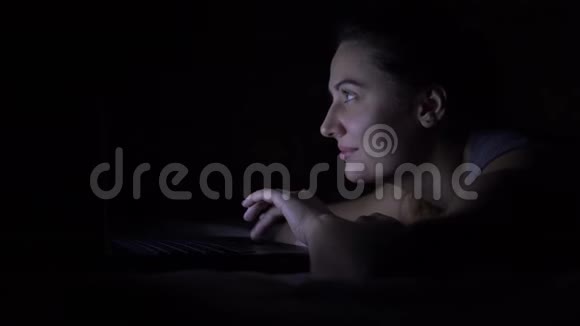 幸福的年轻女人躺在床上在黑暗的房间里用笔记本电脑视频的预览图
