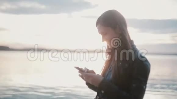 年轻漂亮的女人站在海岸上使用智能手机沙滩上的女孩摸着屏幕微笑视频的预览图