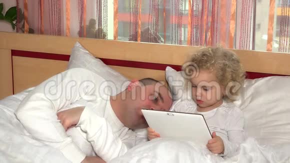 可爱的蹒跚学步的女孩用平板电脑唤醒熟睡的爸爸在床上视频的预览图