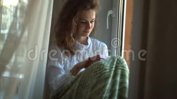 女孩穿着温暖的毯子在窗台上和一只猫喝咖啡一份礼物视频的预览图